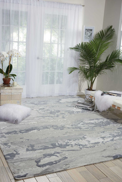 Soft gray and white abstract area rug on pale wood floor with sheer white curtains, palm plant, orchid and fluffy bench.
