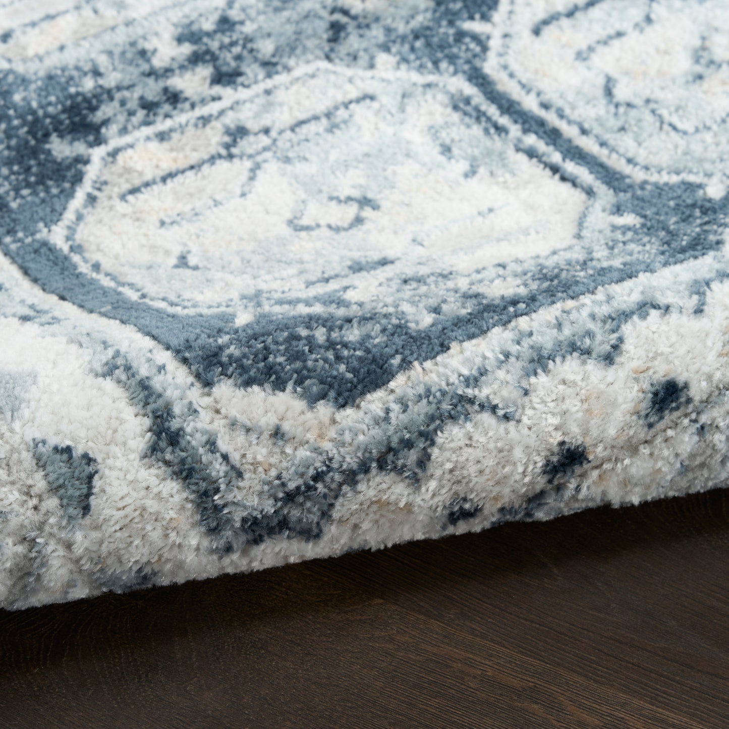 Blue-gray and ivory shag rug with geometric medallion pattern and plush pile on dark wood floor.