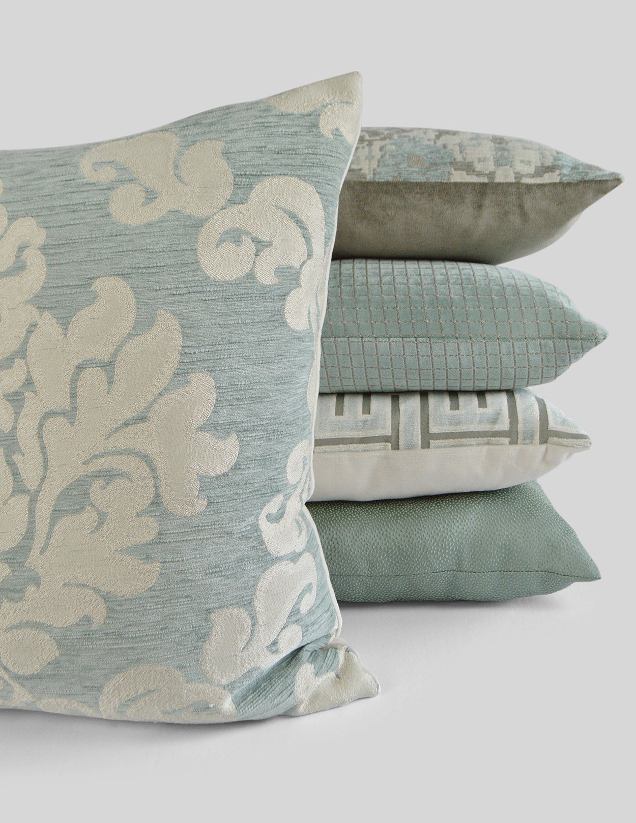Stack of decorative square pillows; front light-blue geometric cushion with cream pattern, behind mint, teal ribbed, and beige geometric cushions.