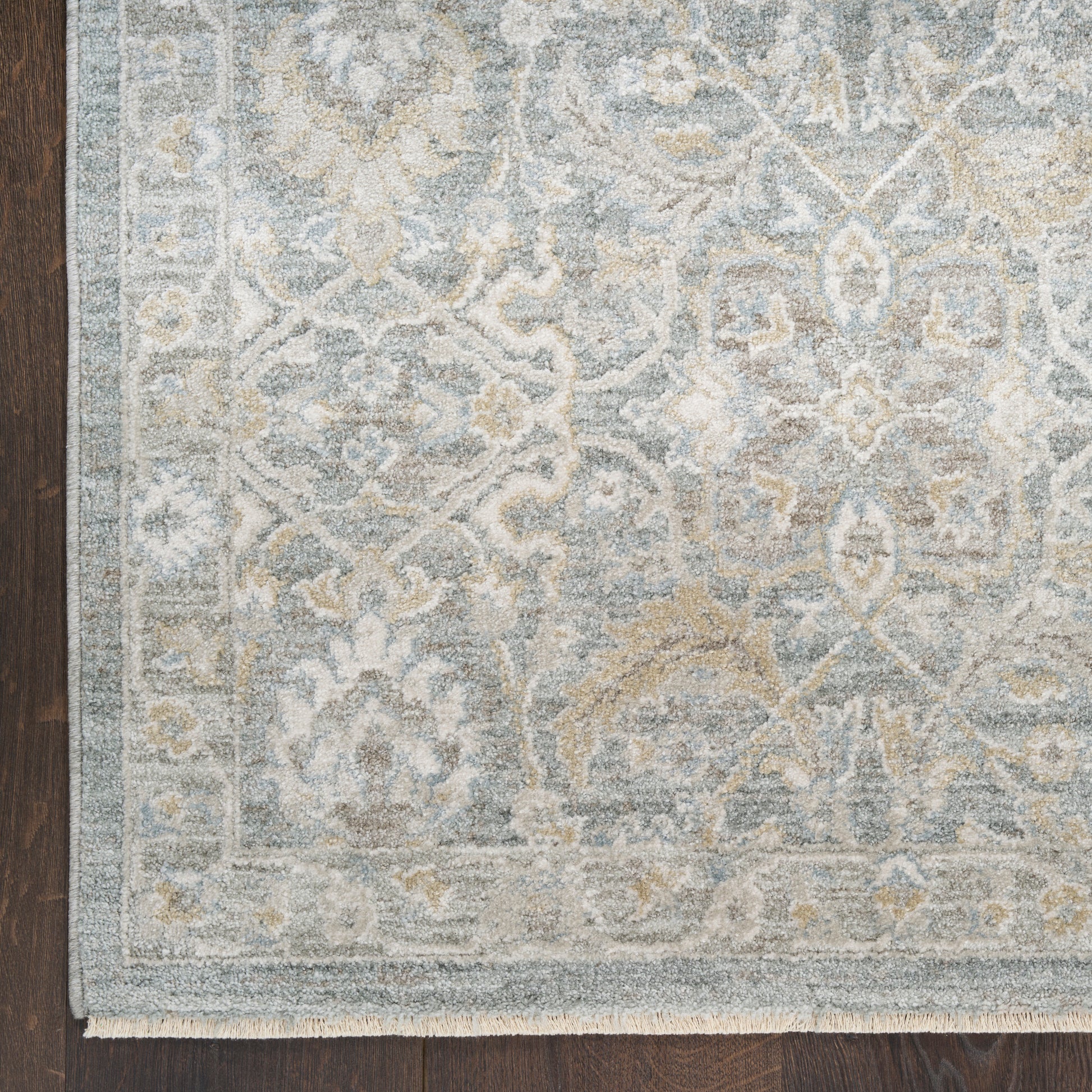 Light blue-gray oriental rug with ivory floral pattern and border, fringed edge on dark wood floor.