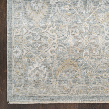 Light blue-gray oriental rug with ivory floral pattern and border, fringed edge on dark wood floor.