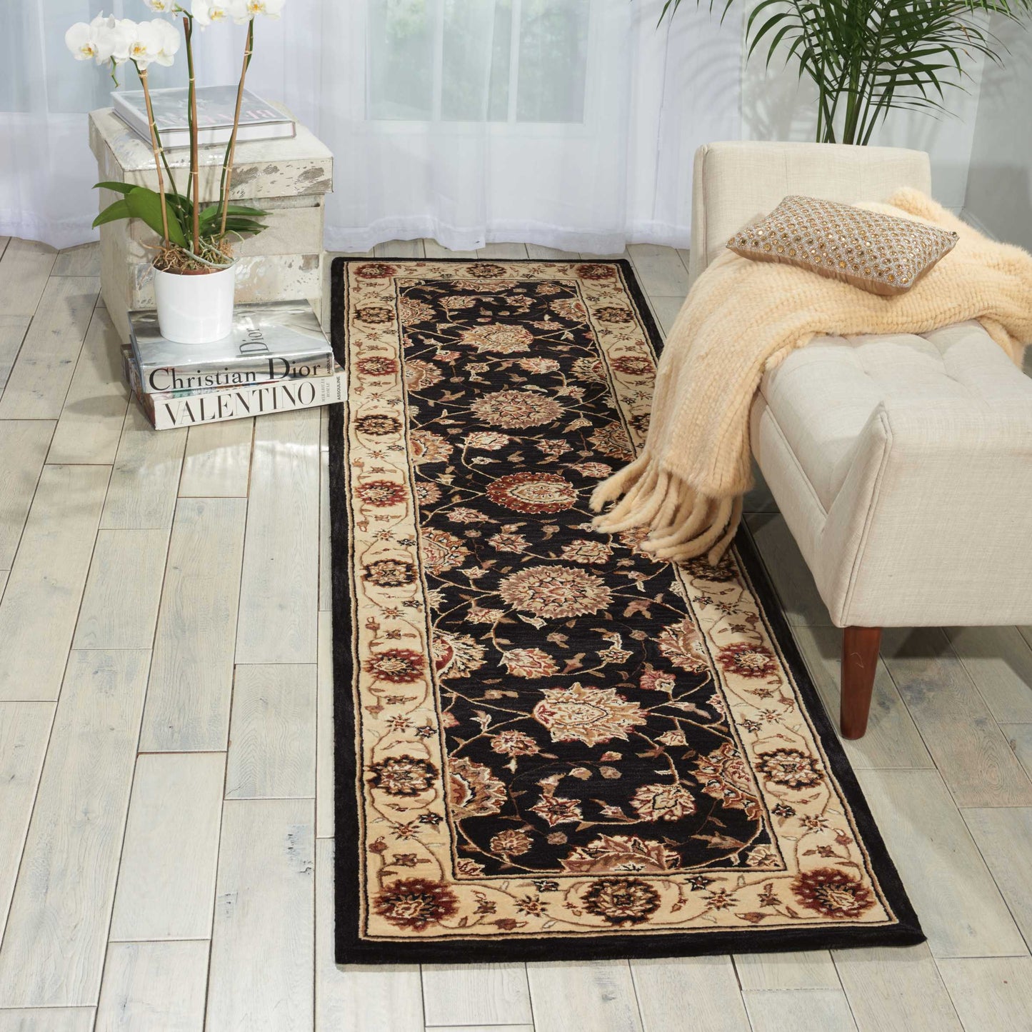 Long oriental rug runner with black center & beige floral border beside a beige sofa, with a potted orchid & fashion books.