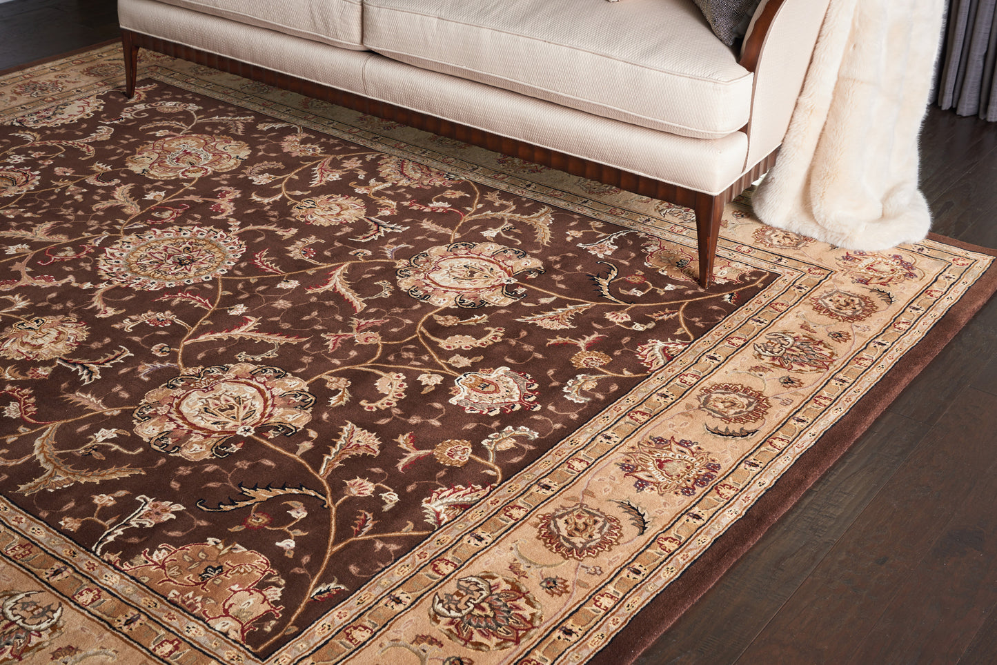 Oriental area rug with dark brown field, cream-gold border and floral motifs, under a beige sofa on dark wood floor.