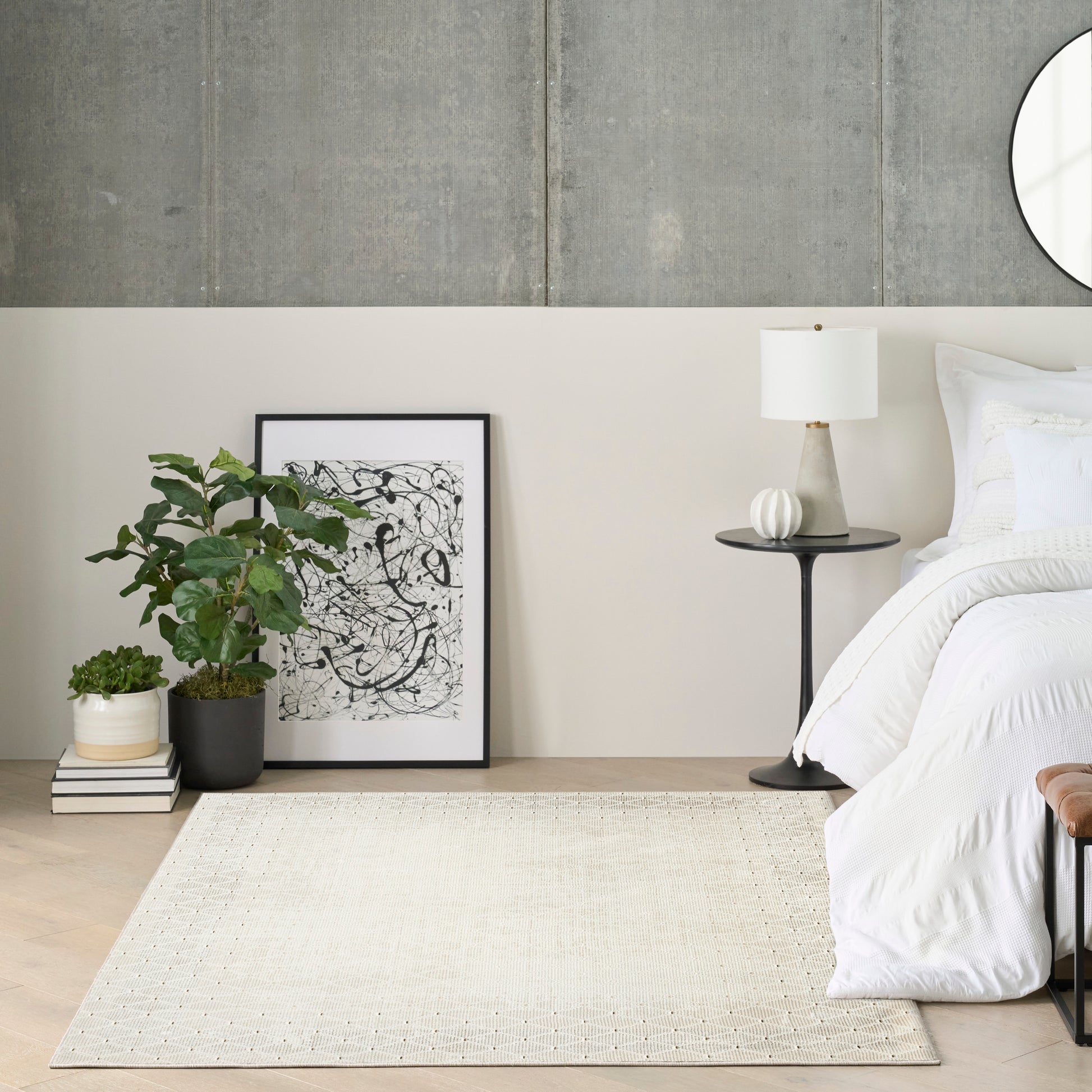 Neutral bedroom with white bedding, black-framed abstract art, potted plant, beige rug, and round side table with lamp.