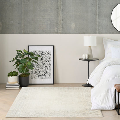 Neutral bedroom with white bedding, black-framed abstract art, potted plant, beige rug, and round side table with lamp.