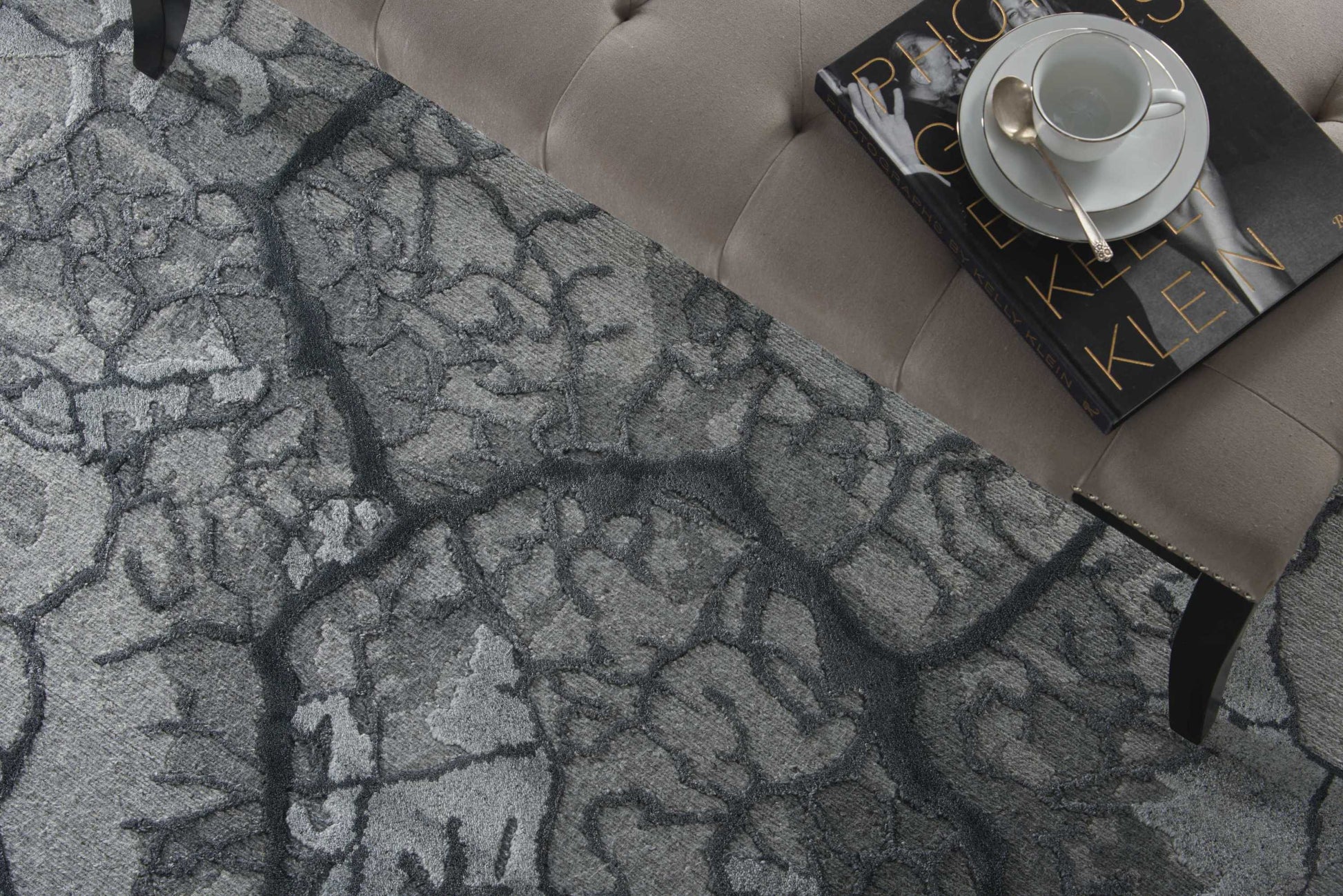 Grey cracked-pattern area rug under a tufted taupe sofa; a magazine with a teacup and saucer rests on top.