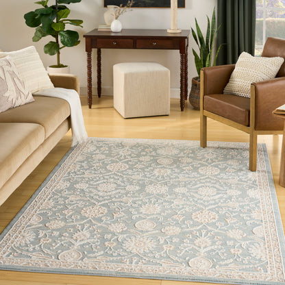 Living room scene with light-blue ornate floral area rug, beige sofa, brown armchair, cream ottoman, wooden console table and plants.