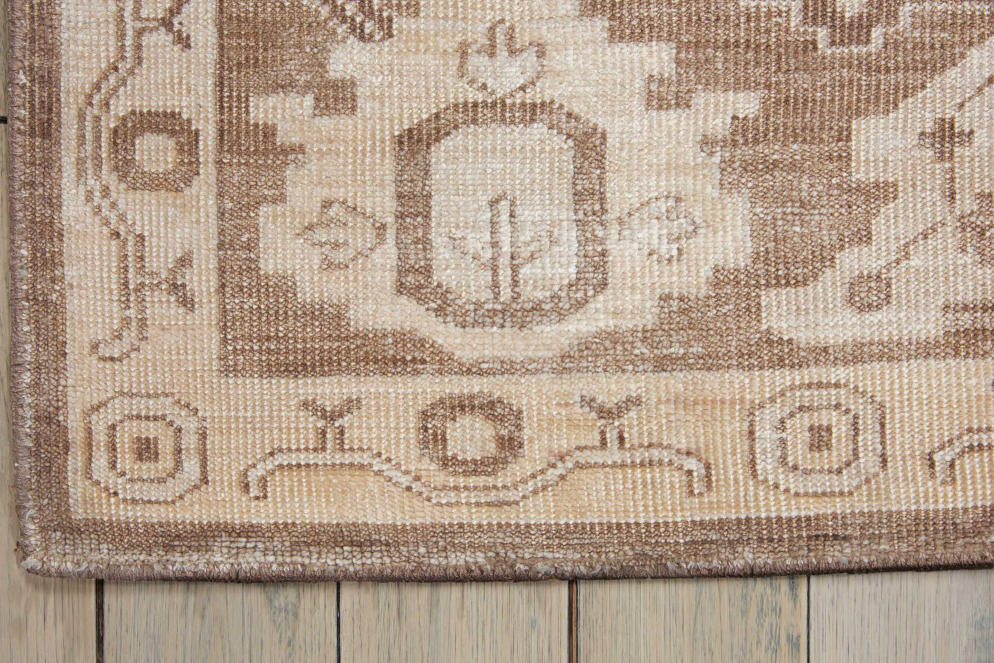 Neutral beige-brown woven rug with geometric medallion and border, close-up corner on wooden floor.