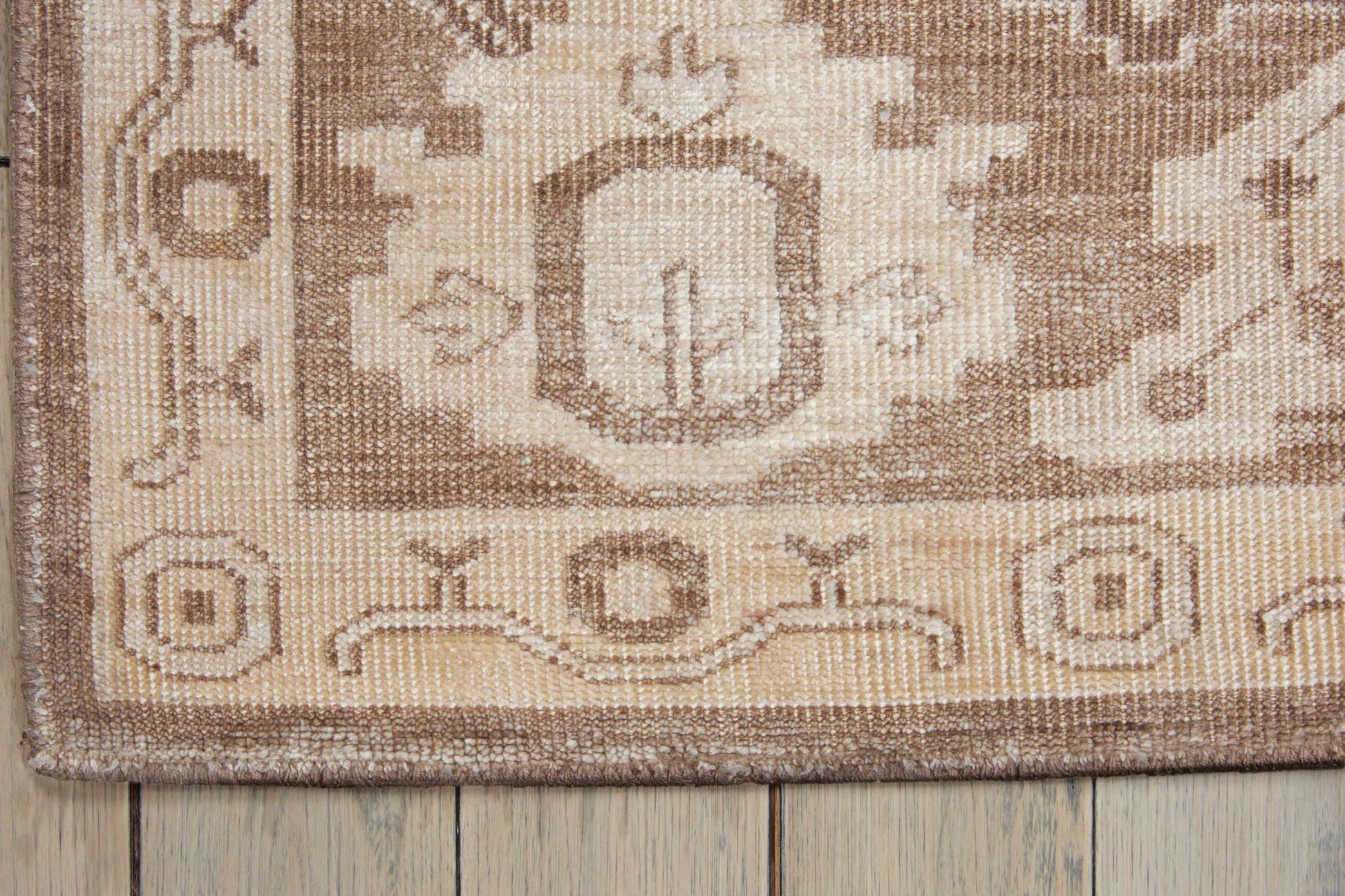 Neutral beige-brown woven rug with geometric medallion and border, close-up corner on wooden floor.