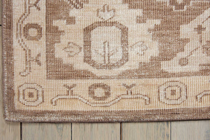 Neutral beige-brown woven rug with geometric medallion and border, close-up corner on wooden floor.