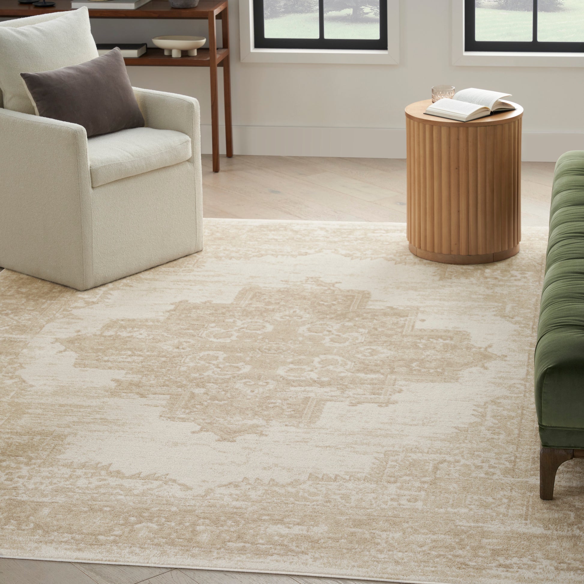 Cream armchair with taupe pillow, round slatted wood side table, open book and glass, beige patterned rug, green sofa edge.