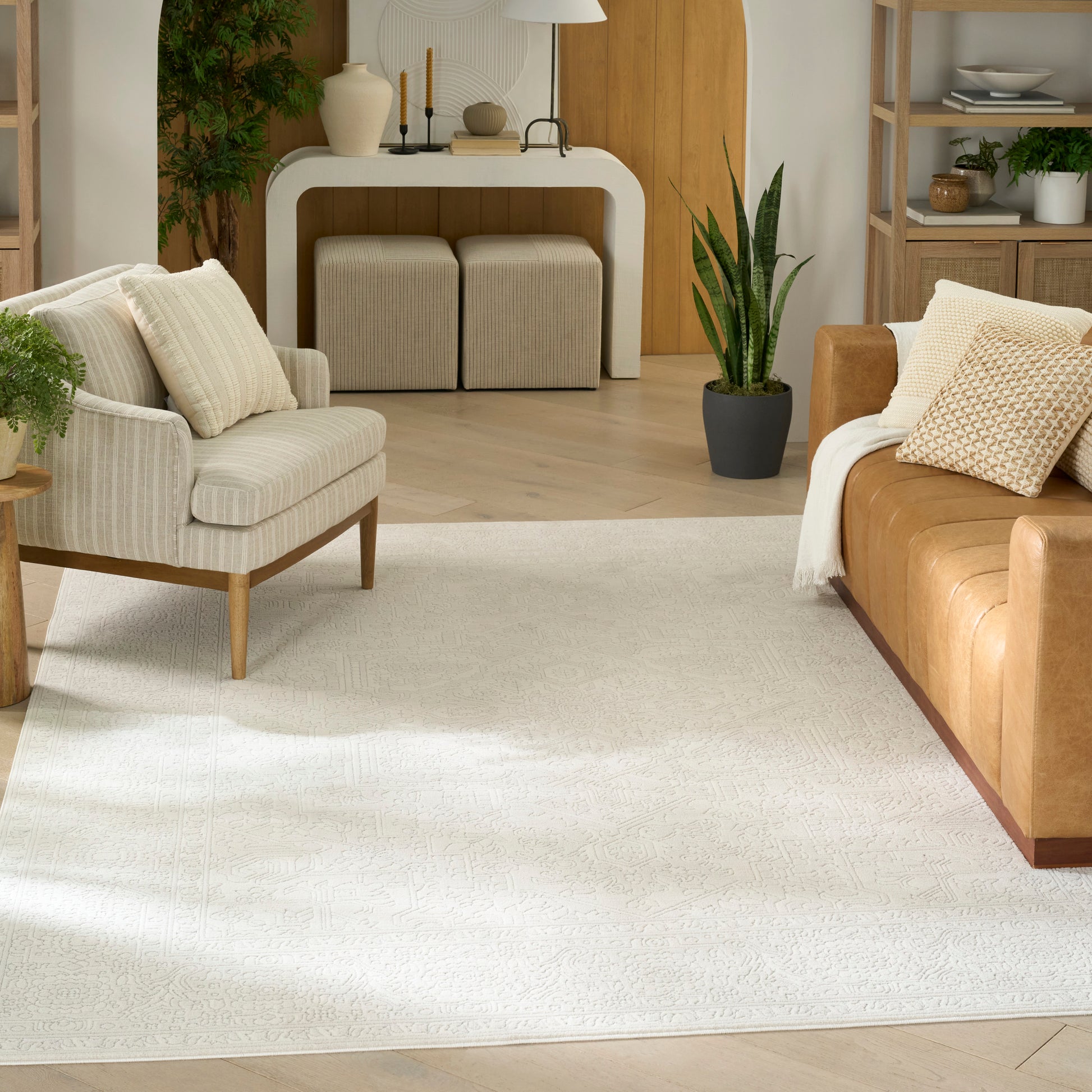 Neutral living room with cream area rug, beige upholstered armchair, tan leather sofa, light wood furniture, and green plants.