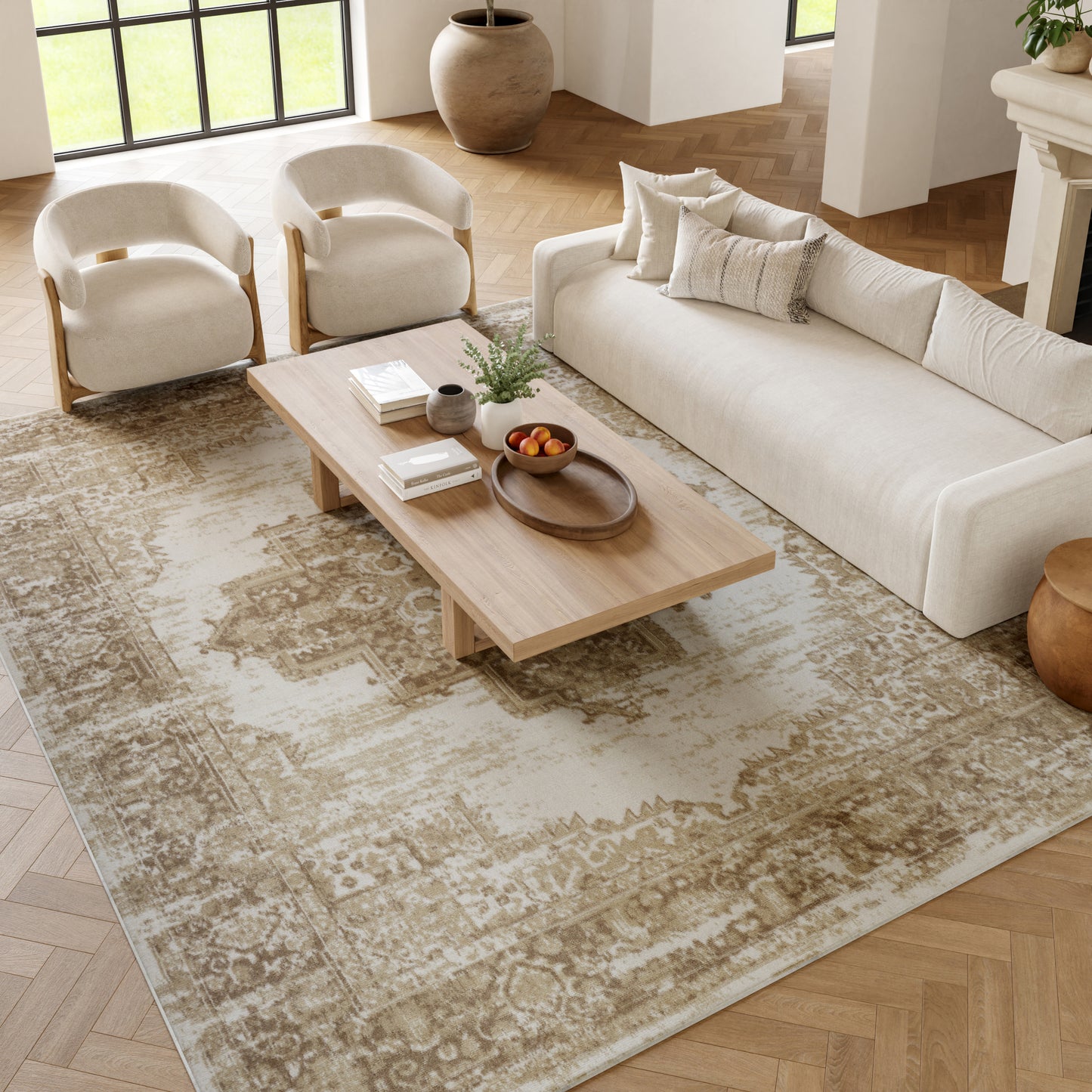Neutral living room with beige sofa, two cream curved armchairs, light wood coffee table with plant & fruit bowl, vintage beige rug.