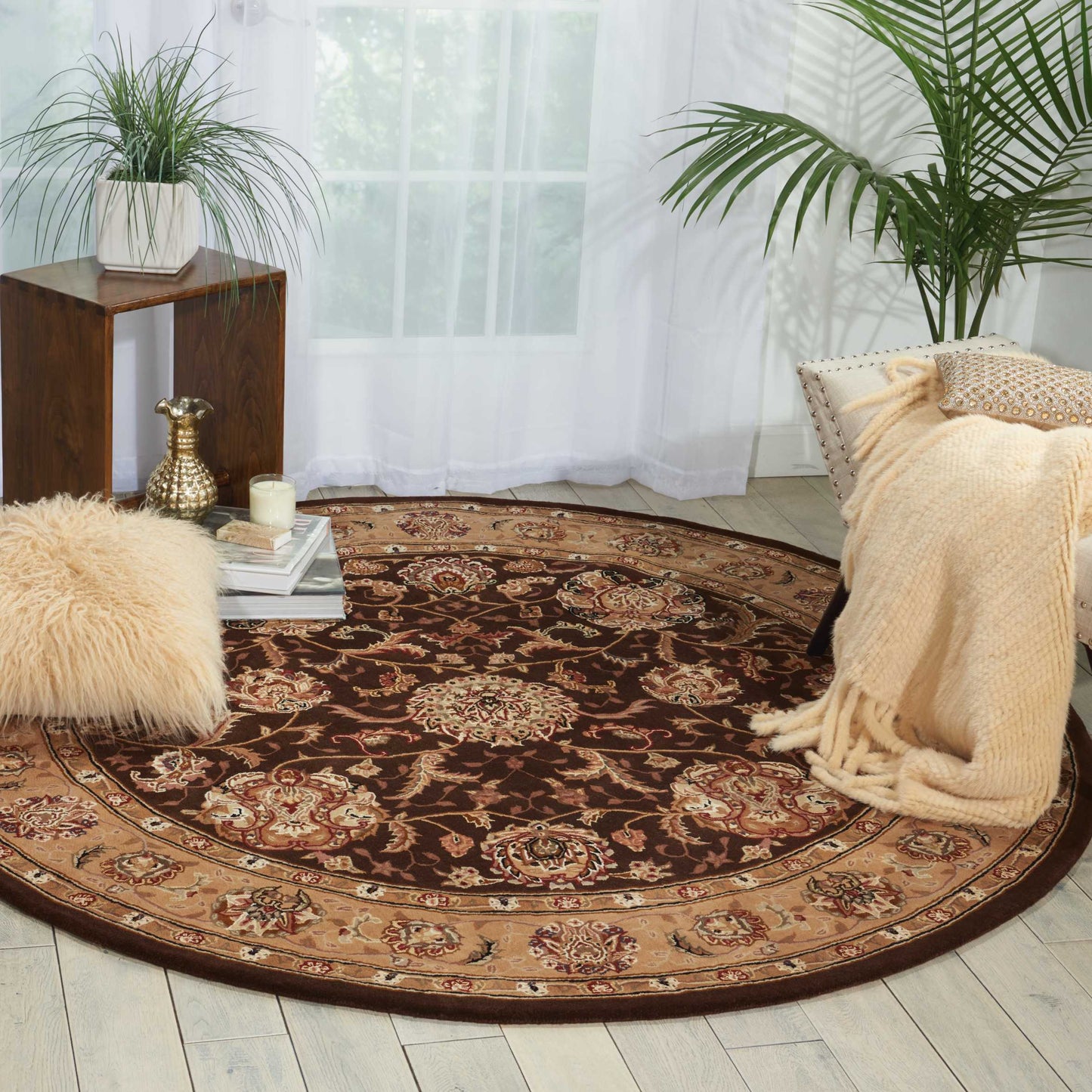Round brown oriental rug with floral border in a bright living room, with a cream blanket and plush ottoman.