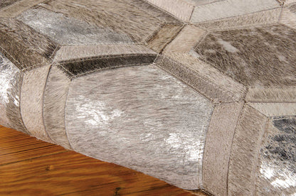 Silver-gray patched cowhide ottoman with geometric hexagon panels and stitched seams on wooden floor.