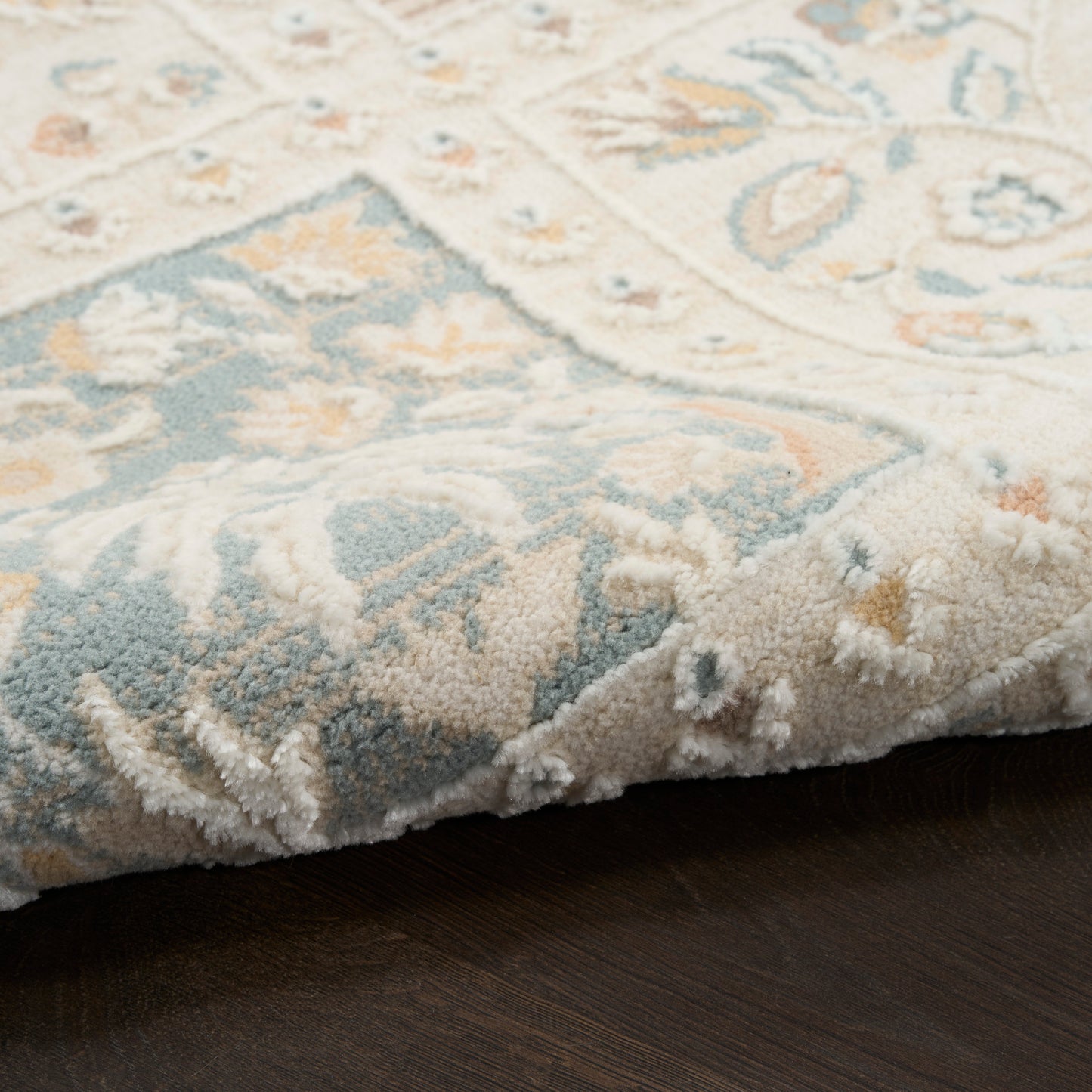 Soft area rug with pale blue, cream and beige floral pattern, slightly folded over on dark wood floor.