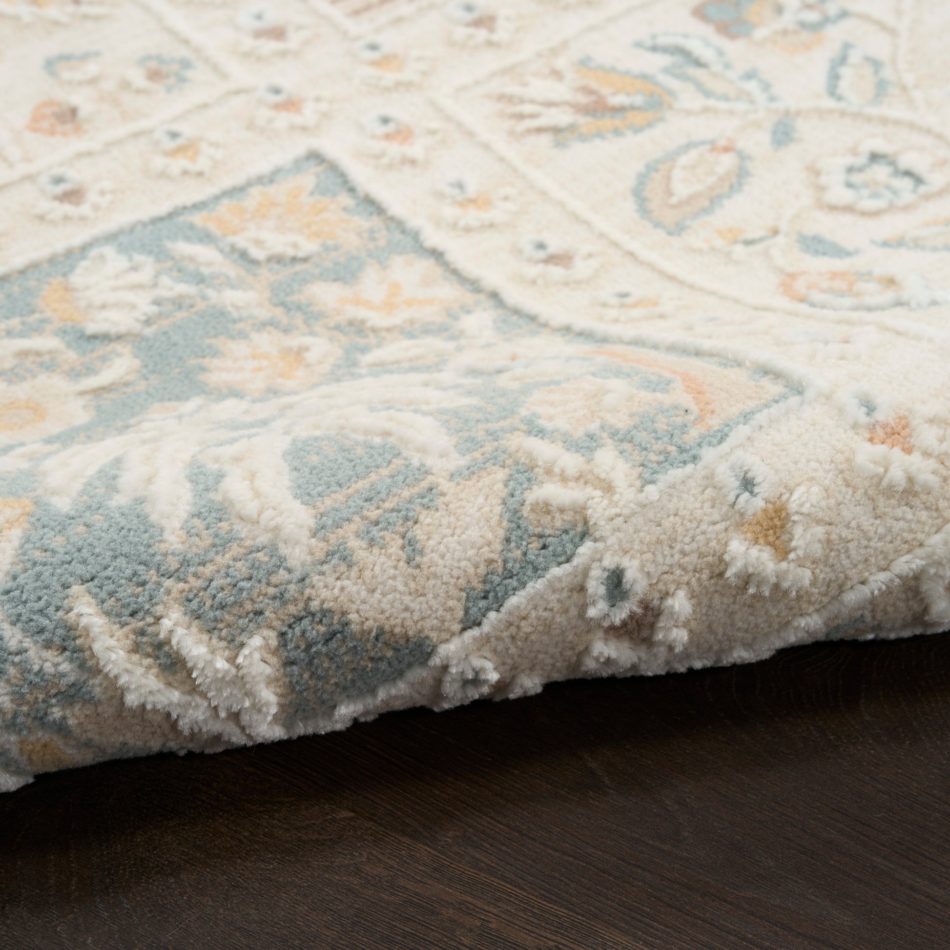 Soft area rug with pale blue, cream and beige floral pattern, slightly folded over on dark wood floor.