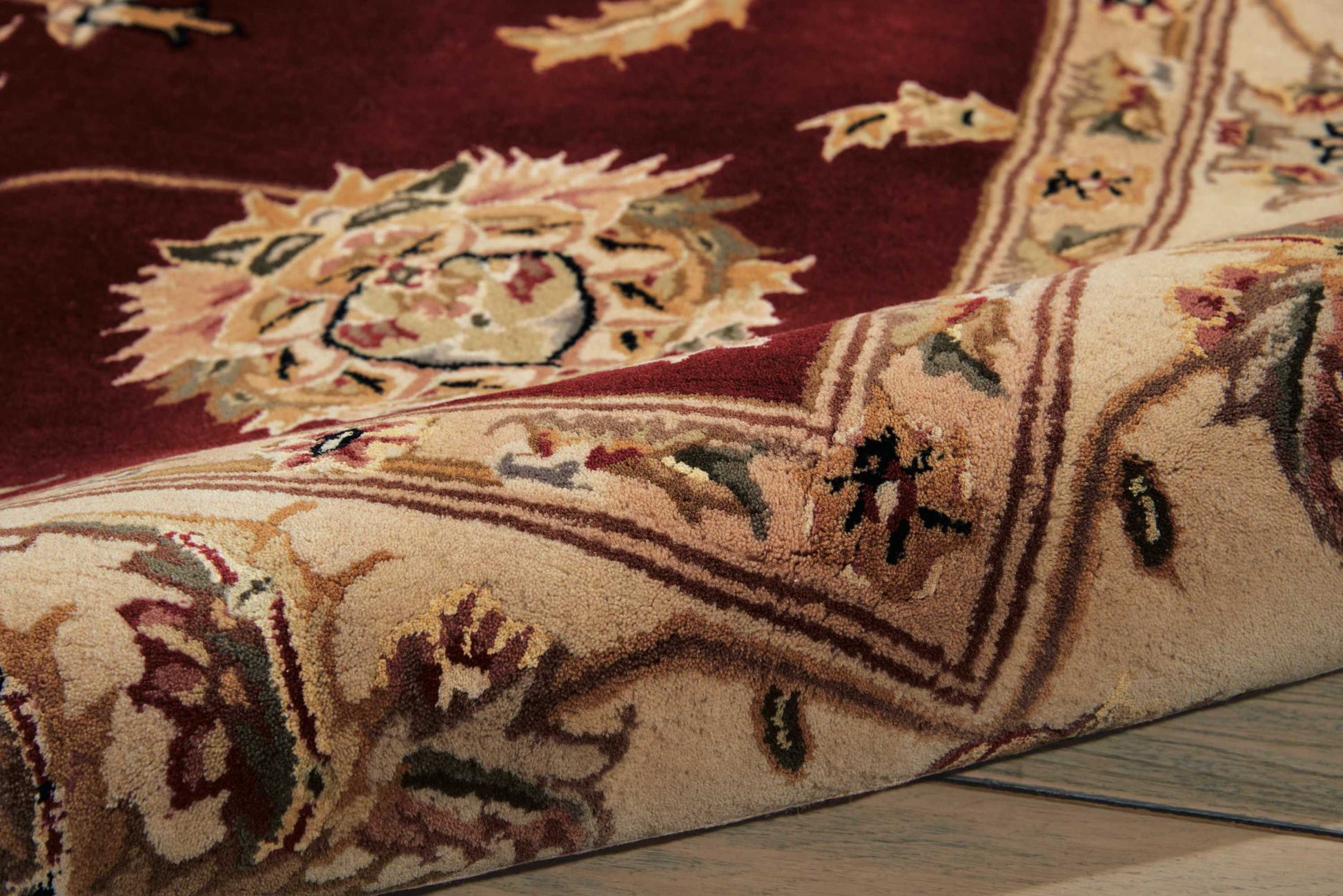 Burgundy oriental rug with cream floral border and medallion pattern, soft pile; edge folded over on wood floor.