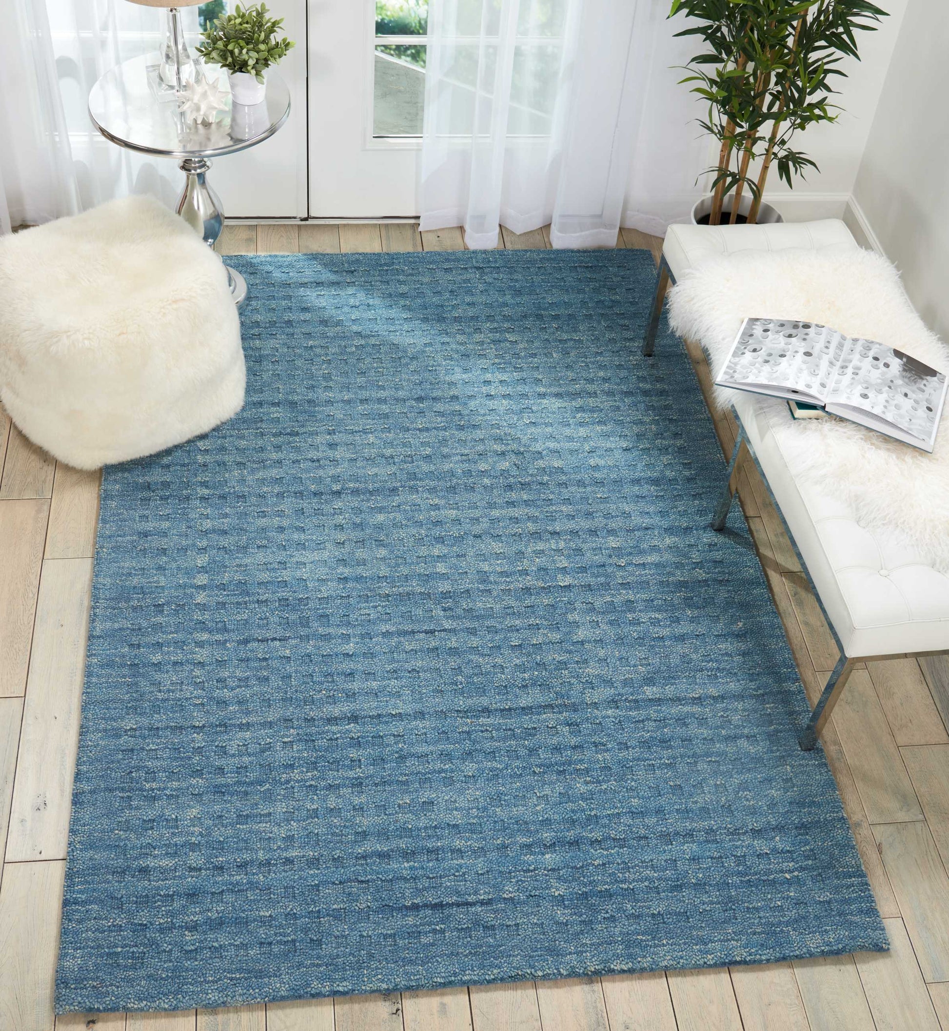 Blue textured flat-weave rug in a bright living room with a white tufted bench and fluffy white pouf.