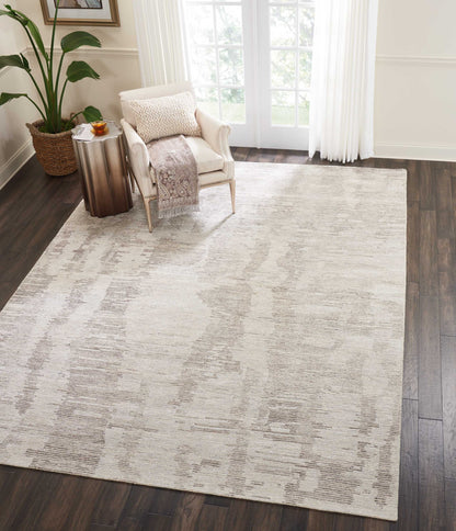 Neutral beige-gray abstract area rug on dark wood floor in bright living room with cream armchair and sheer curtains.