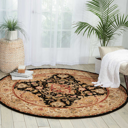 Round Persian-style rug with black center and red-beige border on light wood floor, with plants, books, and a chair.