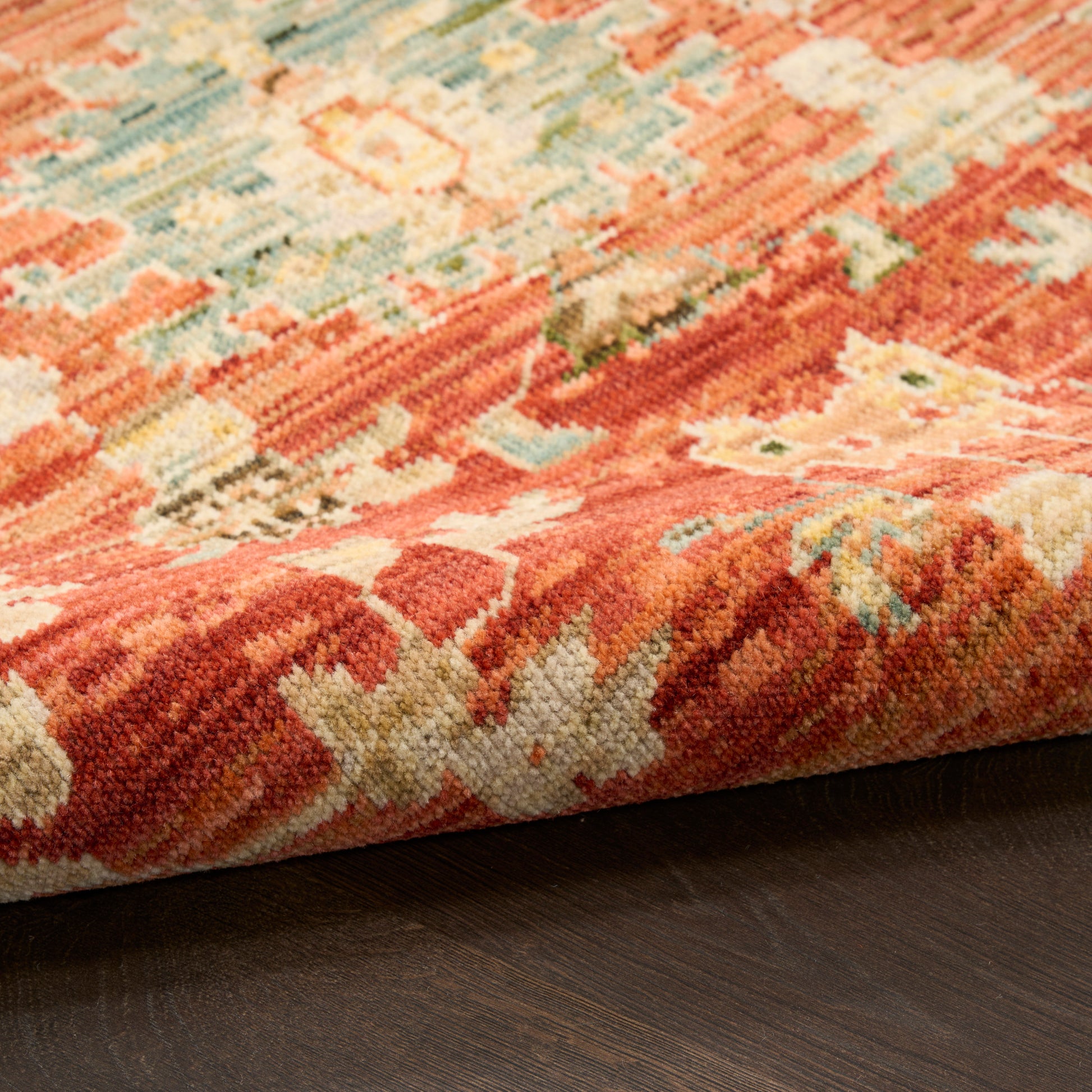 Close-up of a vibrant oriental rug with red, coral, teal and cream motifs, folded edge on dark wood floor.