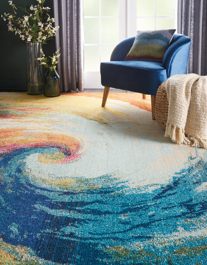 Blue velvet armchair beside a colorful swirl rug, beige knit throw, and branch vase by the window.