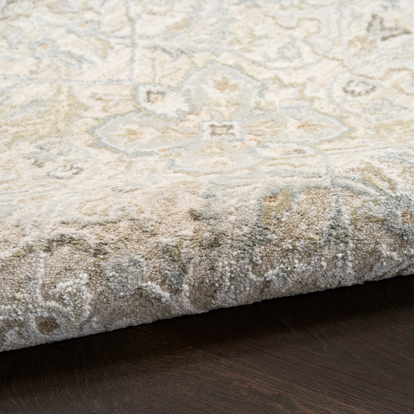 Close-up of a cream oriental rug with grey-blue floral pattern, folded edge on a dark wooden floor.