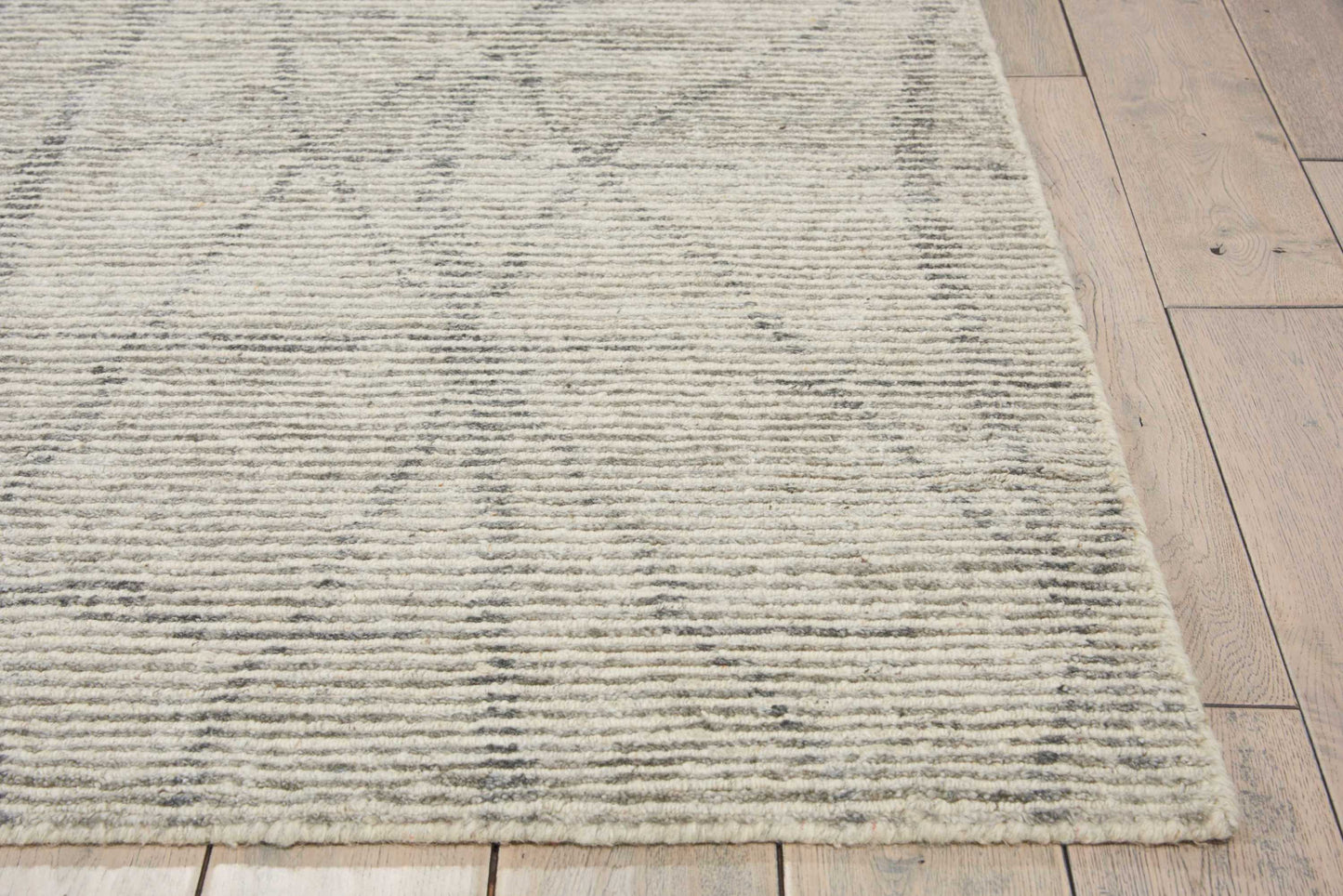 Light gray-beige area rug with subtle diamond pattern, ribbed texture and fringed edge on a wooden floor.