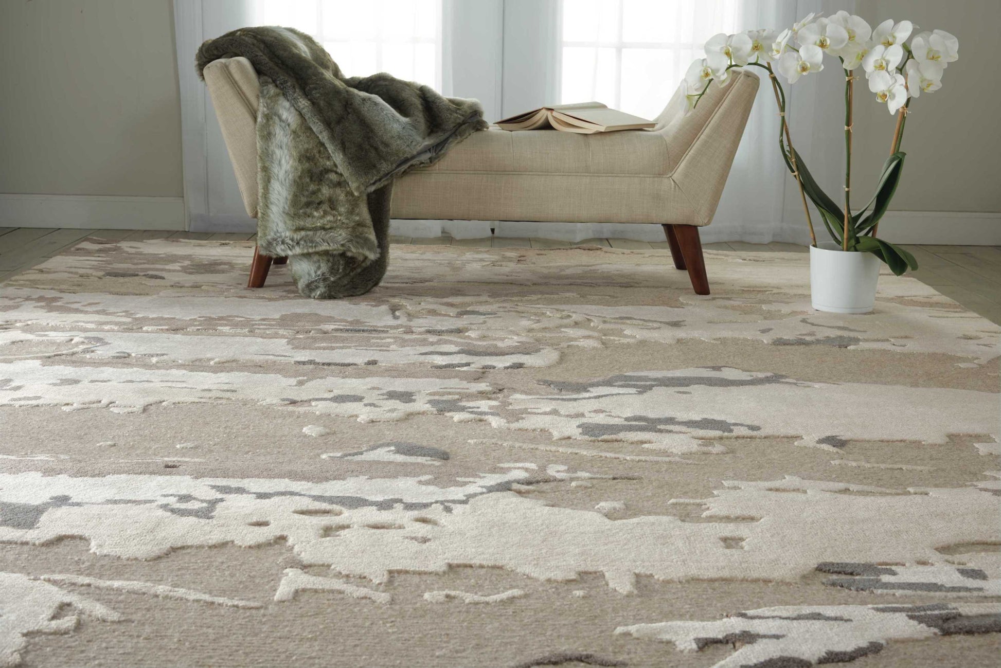 Beige chaise lounge with wooden legs, gray faux fur throw, open book, on a neutral abstract rug; white orchid in a pot.
