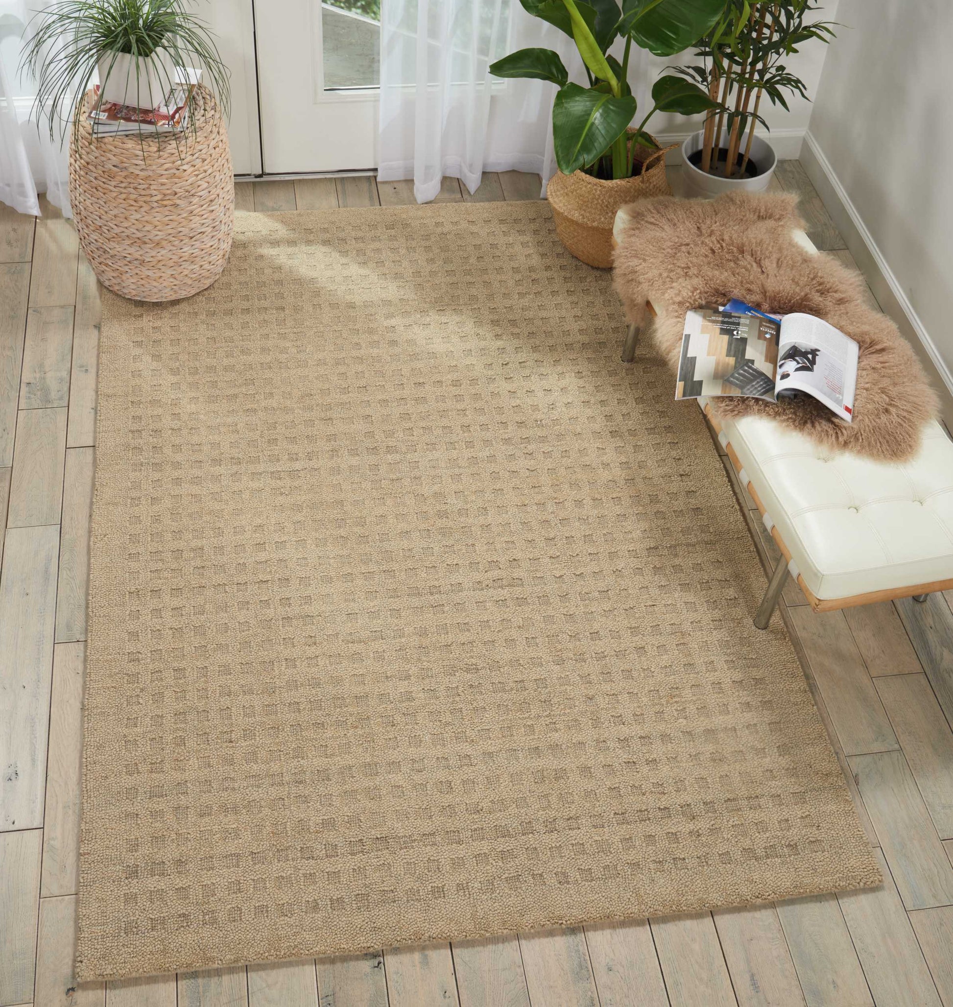 Beige woven jute rug with subtle grid texture in a sunlit living room with plants and a bench topped by a faux fur throw.