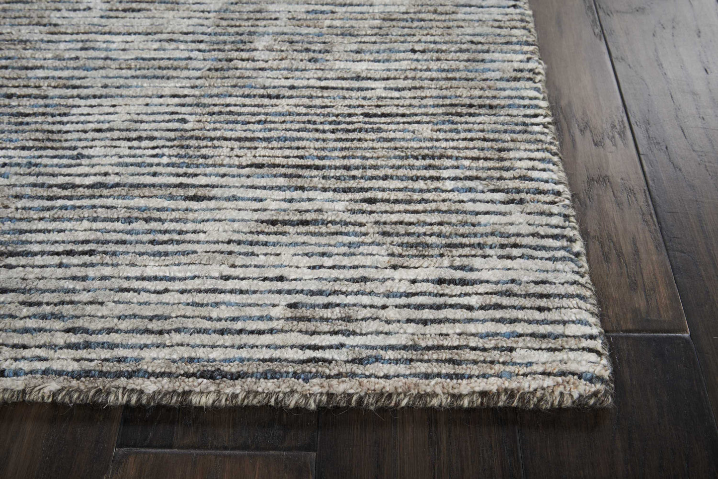Textured blue-gray striped rug with fringed edge on dark hardwood floor.