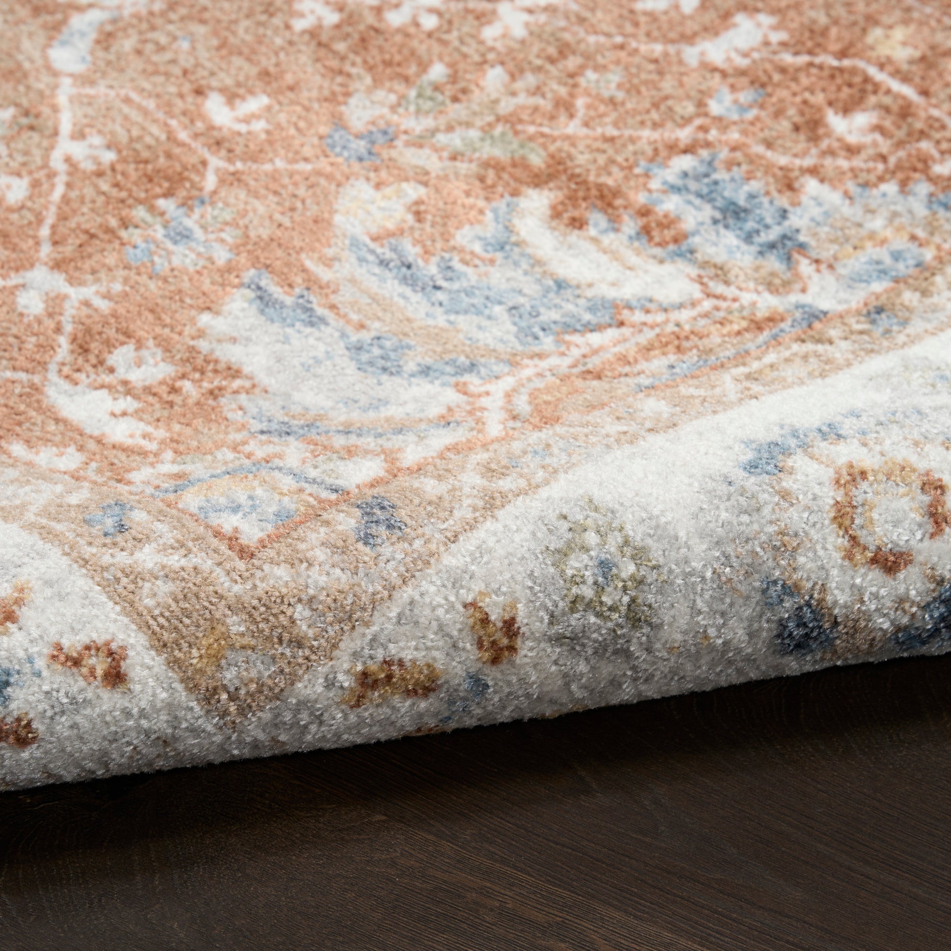 Close-up of an oriental rug with orange, blue and ivory floral motifs, its edge slightly curled on dark wood.