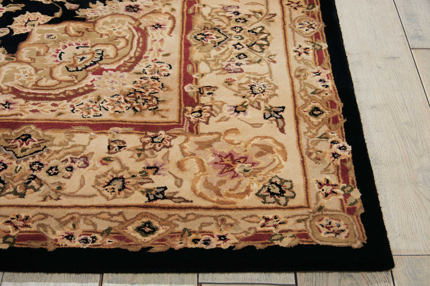 Beige ornate rug with floral patterns, gold border and black edge on light wood floor.