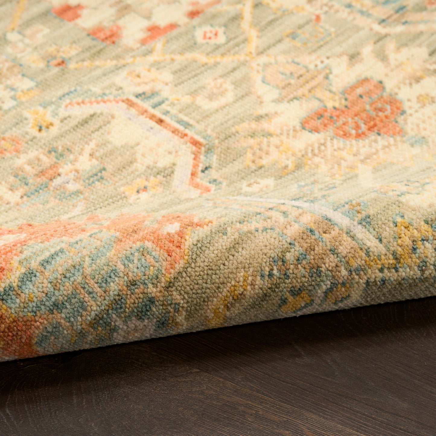 Close-up of a woven pastel rug with geometric motifs in blue, peach and beige; edge visible on dark wood floor.