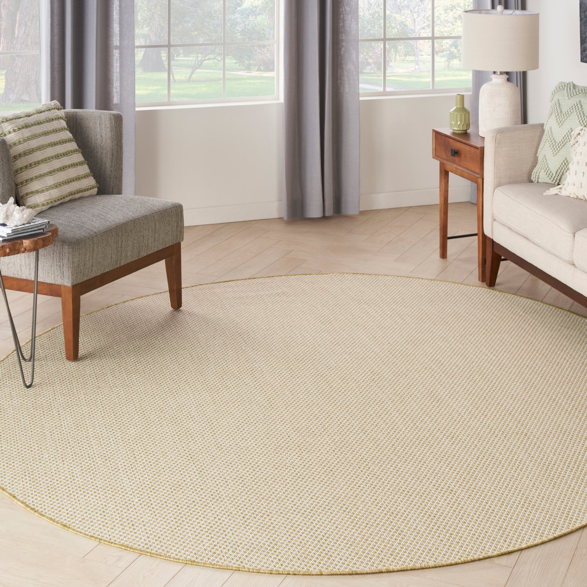 Round 8' natural jute rug in a light living room with gray armchairs, wooden side tables and curtains.