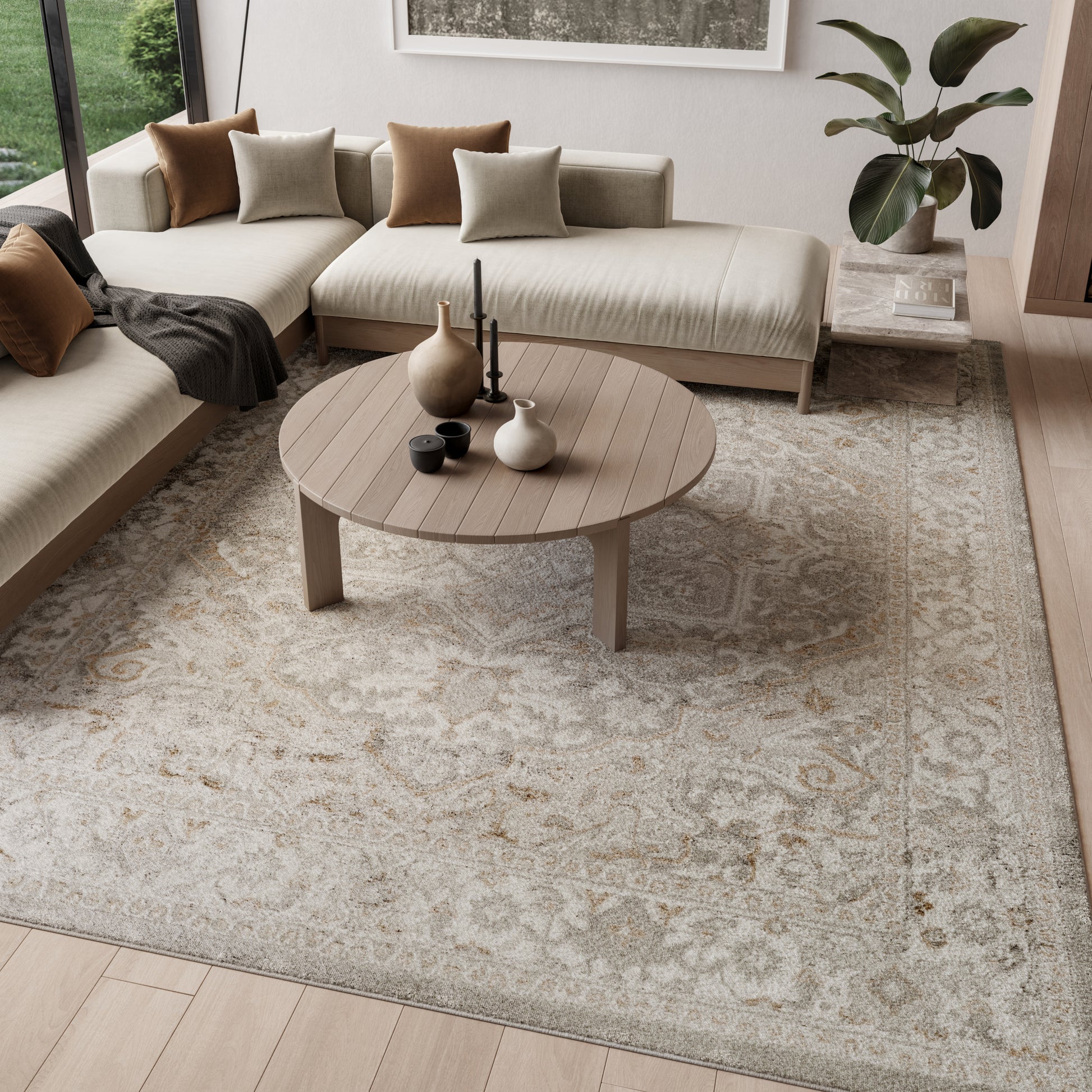Light beige sectional sofa with brown & taupe cushions, round wood coffee table, patterned rug, plant accents & vases.