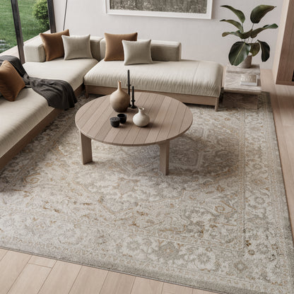 Light beige sectional sofa with brown & taupe cushions, round wood coffee table, patterned rug, plant accents & vases.