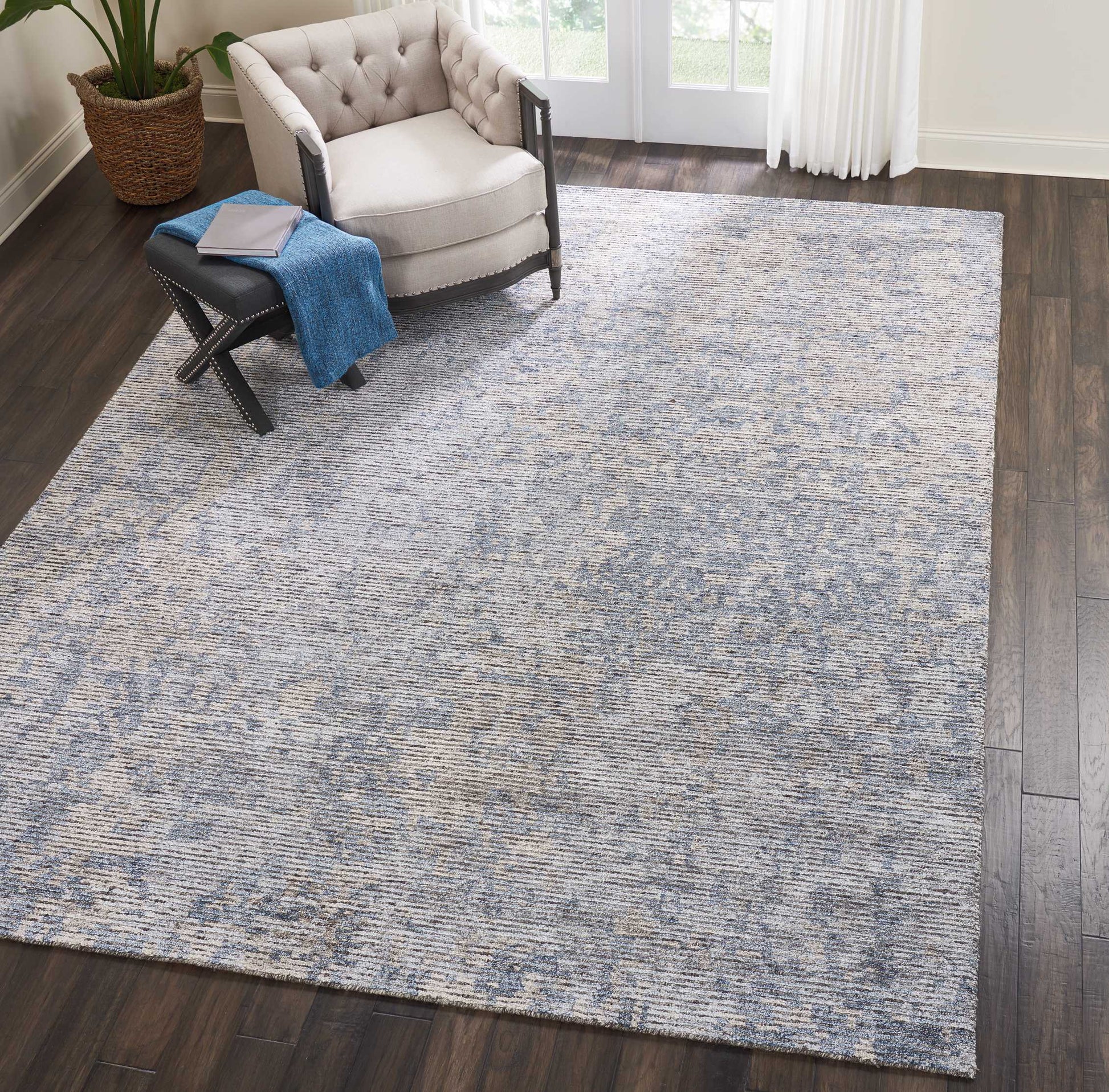 Blue-gray textured rectangular area rug on dark wood floor beside a tufted beige armchair and black ottoman with a blue throw.