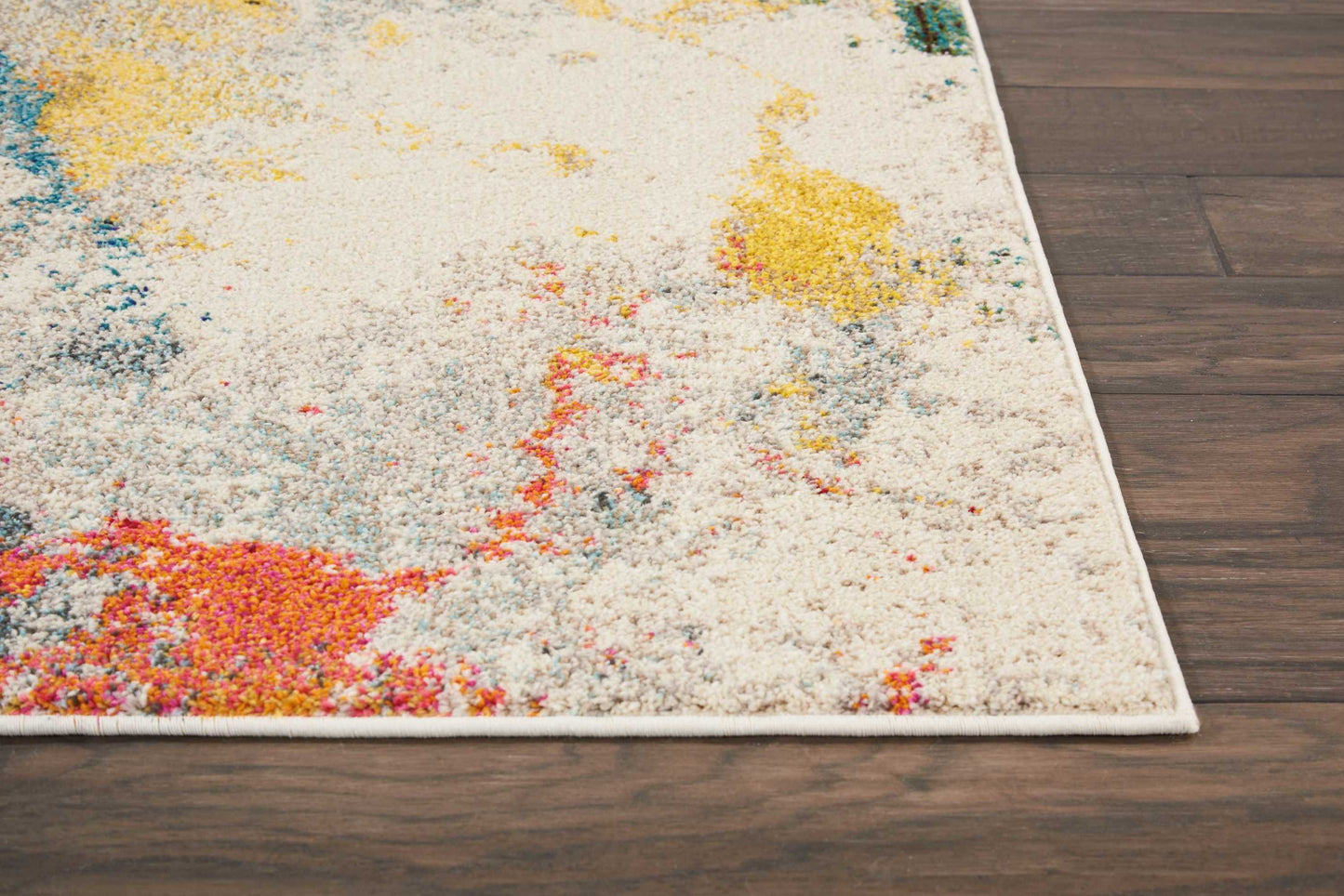 Plush cream abstract area rug with pink, yellow, blue and orange splashes on a dark wood floor; fringed edge.