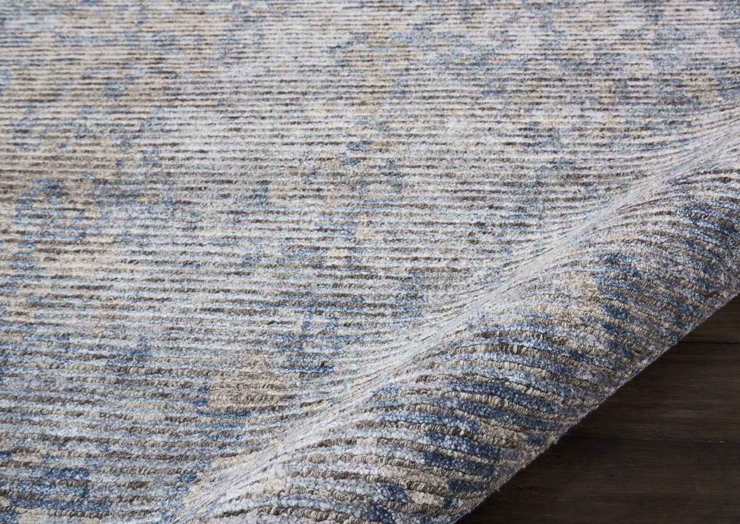 Blue-gray distressed area rug with soft woven texture and subtle pattern; rolled corner on dark wood floor.