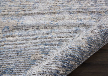 Blue-gray distressed area rug with soft woven texture and subtle pattern; rolled corner on dark wood floor.