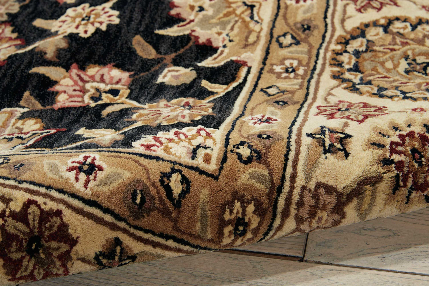 Beige and black oriental rug with ornate floral border and red accents; plush pile, corner folded on wood floor.