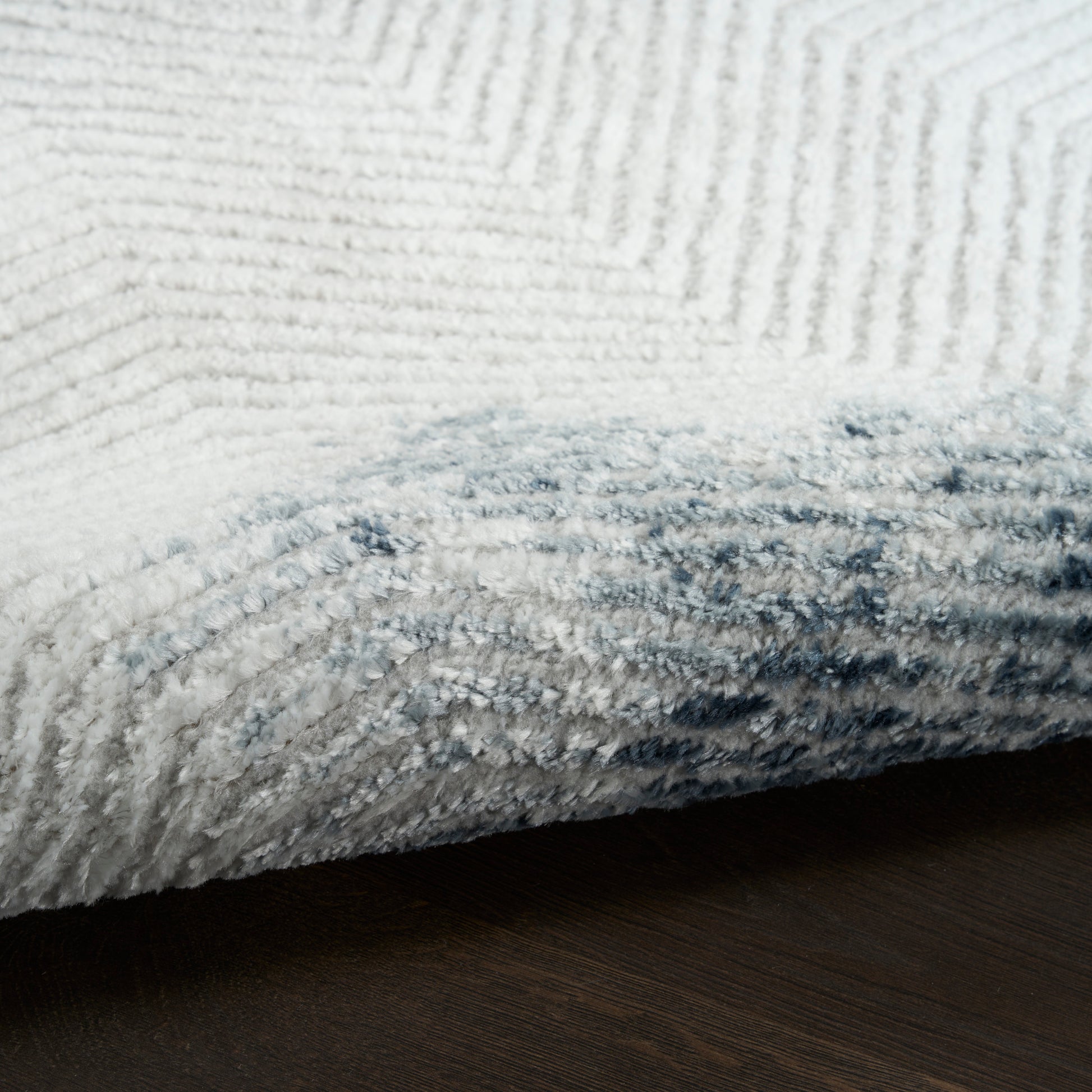 Close-up of white and pale blue shag rug with thick plush pile, rolled edge on dark wood floor.