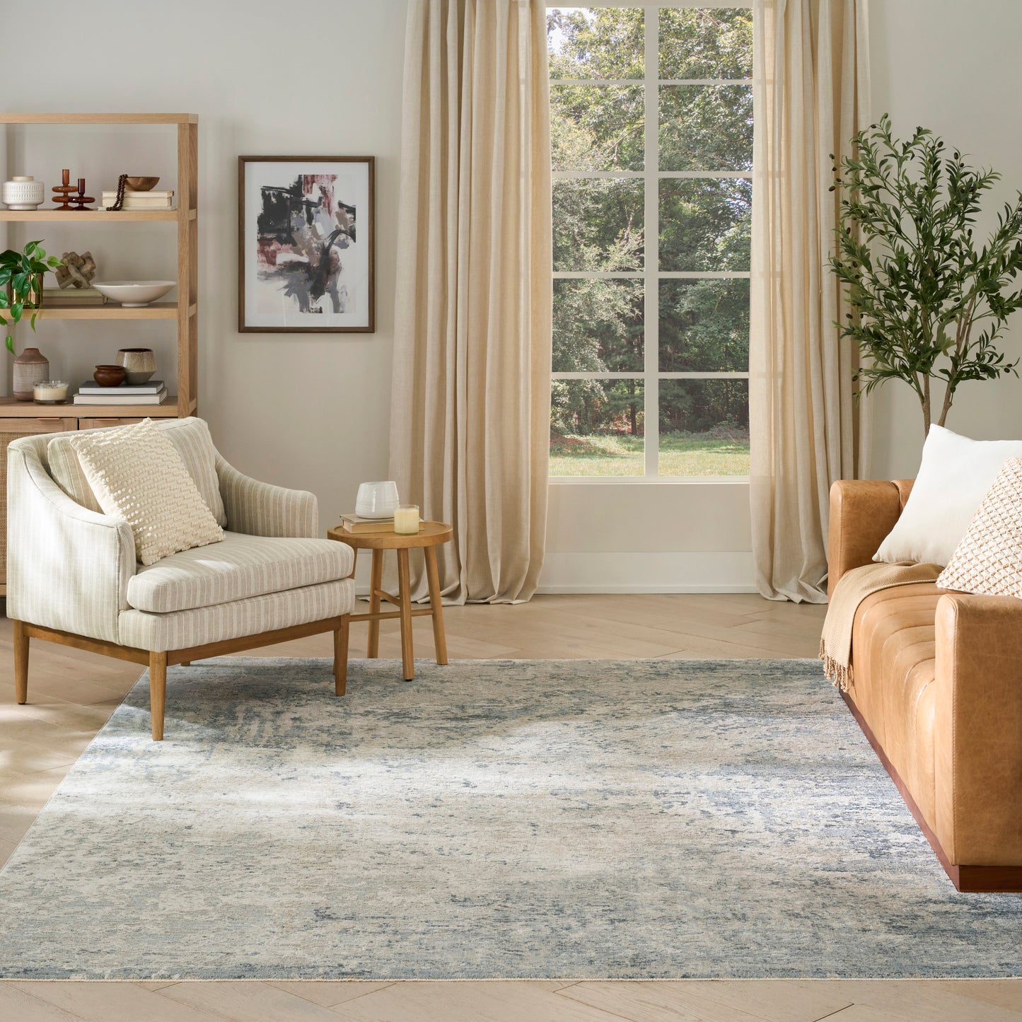 Cozy living room with cream armchair, tan sofa, wooden shelves, abstract art, window with beige curtains, blue rug & plant.