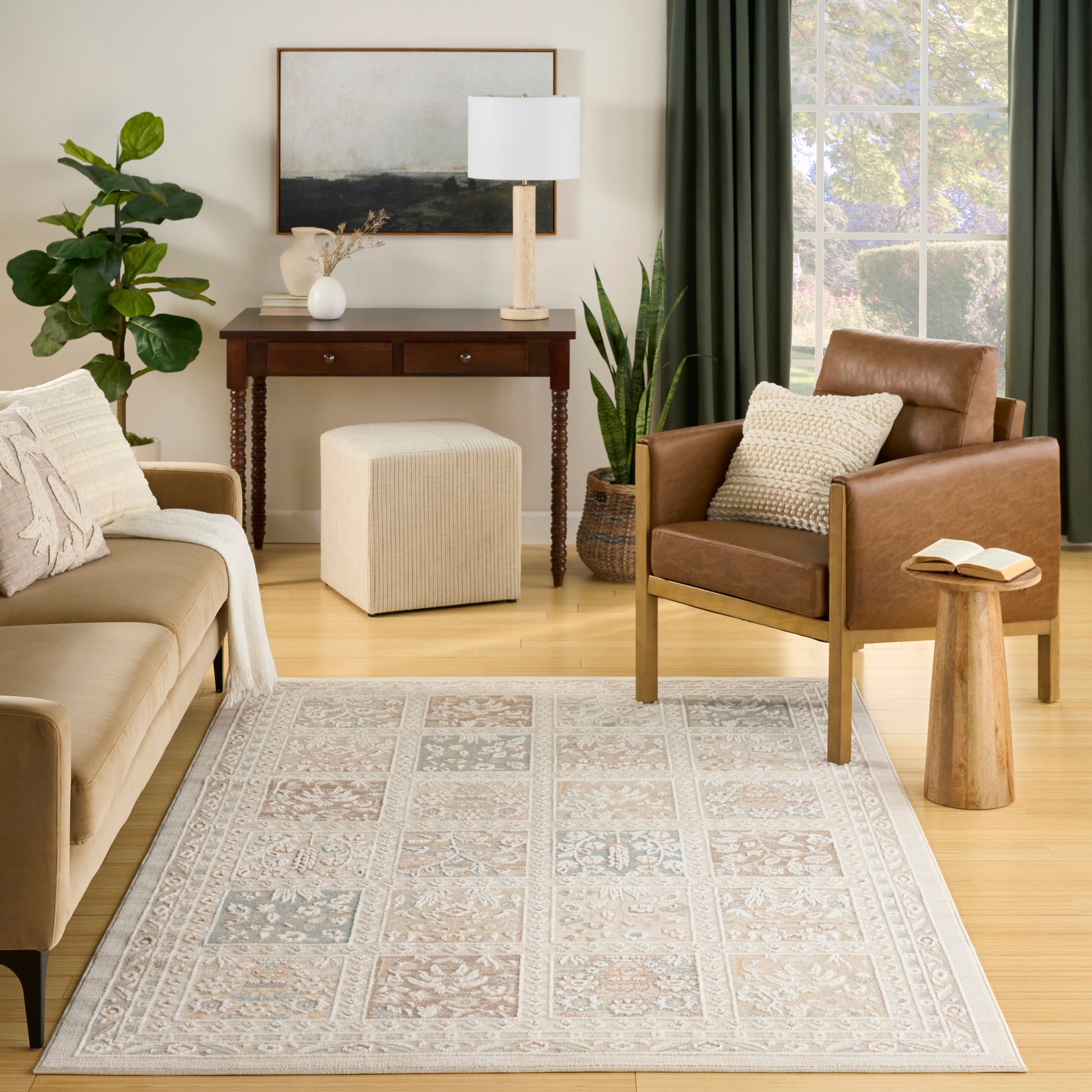 Cozy living room with tan sofa, brown leather chair, pastel patterned rug, wood console, green-curtained window, and plants.