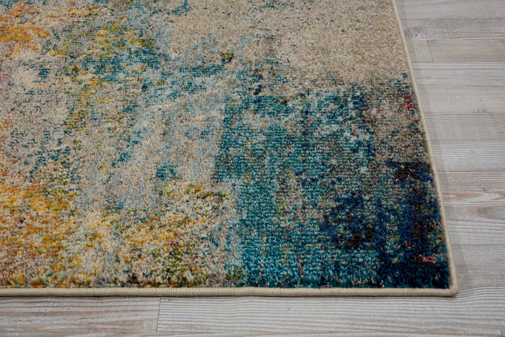 Abstract rectangular rug with beige, blue, teal and yellow tones, distressed pattern, bound edge on light wood floor.