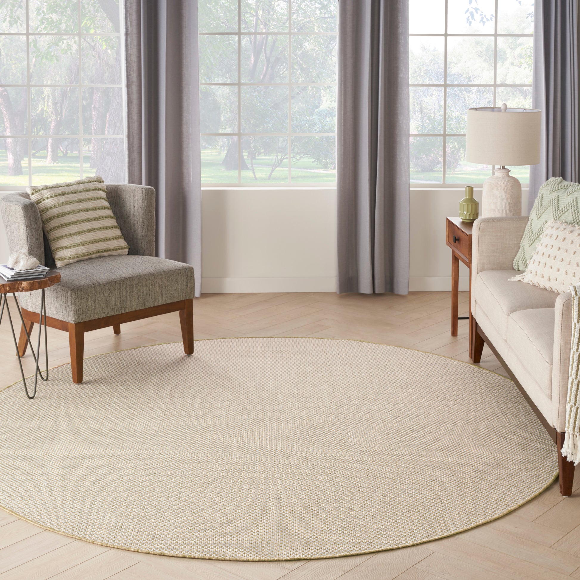 Circular beige woven area rug in a light living room with gray chair, beige sofa, wood end tables, lamp, and large windows.