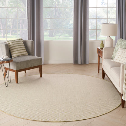 Circular beige woven area rug in a light living room with gray chair, beige sofa, wood end tables, lamp, and large windows.