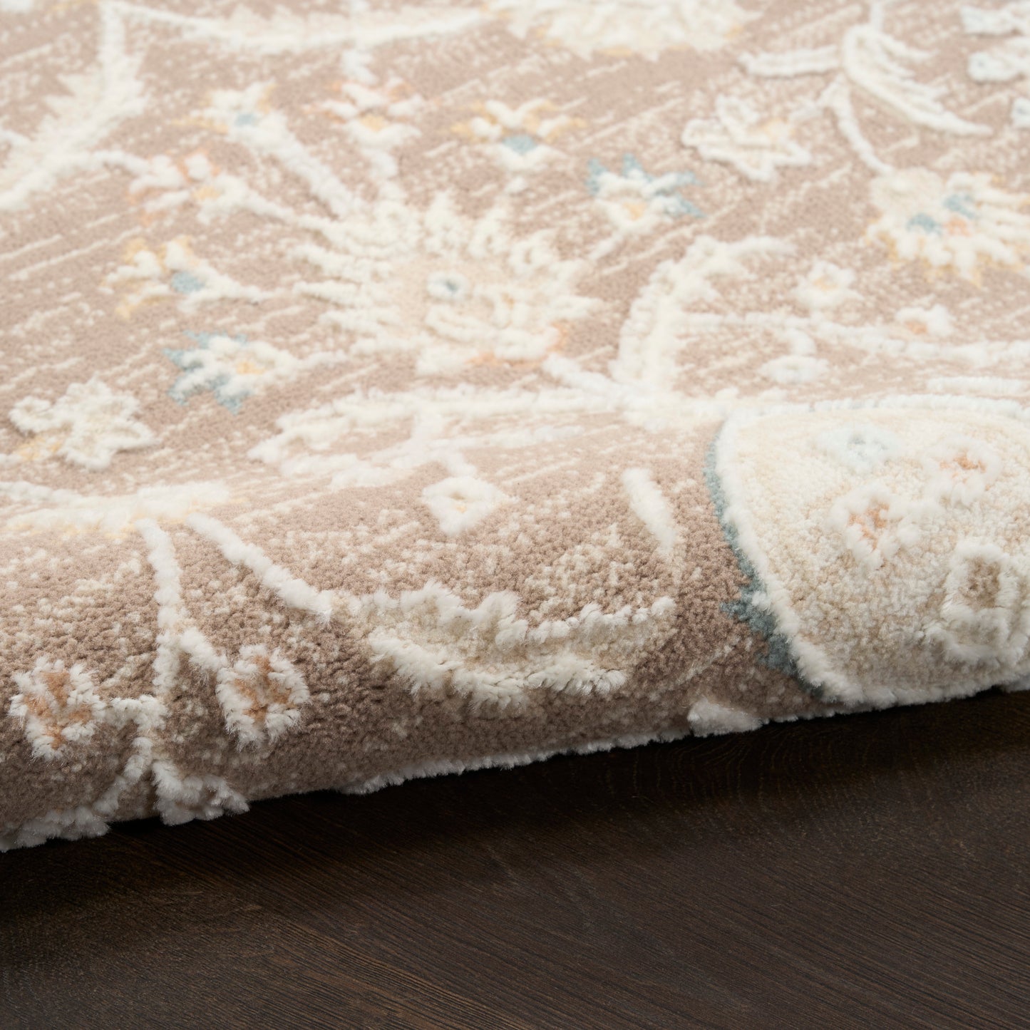 Beige floral area rug with white pile and pale blue accents, rolled edge on dark wooden floor.